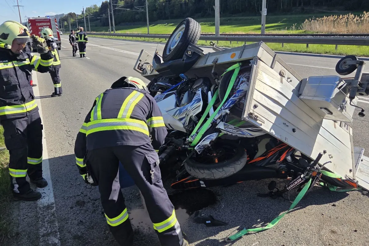 Feuerwehrleute stehen auf einer Landstraße an einem umgestürzten Anhänger, unter dem ein Motorrad zu sehen ist. Die Szenerie ist klar und bei Tageslicht, mit grüner Landschaft im Hintergrund.