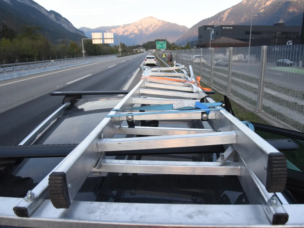 Eine Leiter ist unsicher auf dem Dach eines Fahrzeugs befestigt, das auf einer Autobahn steht. Im Hintergrund sind Berge sichtbar.