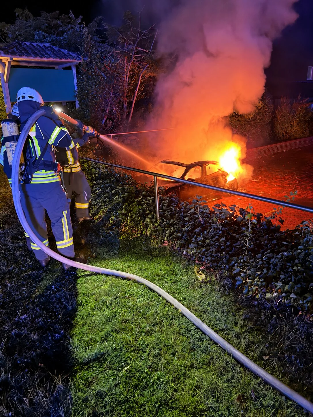 Ein Feuerwehrmann bekämpft bei Dunkelheit einen Fahrzeugbrand mit einem Wasserschlauch. Flammen und Rauch steigen aus dem Auto auf.