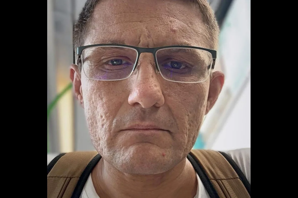 Das Bild zeigt einen Mann mit Brille und einem ernsten Gesichtsausdruck. Im Hintergrund sind unscharfe helle Strukturen erkennbar.