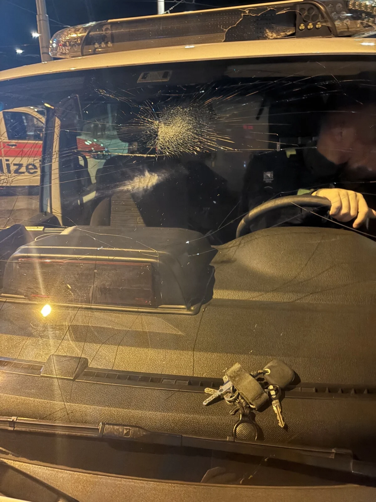 Ein Polizeifahrzeug mit gesplitterter Windschutzscheibe, sichtbar bei Nachtaufnahme. Der Fahrer ist angedeutet erkennbar.