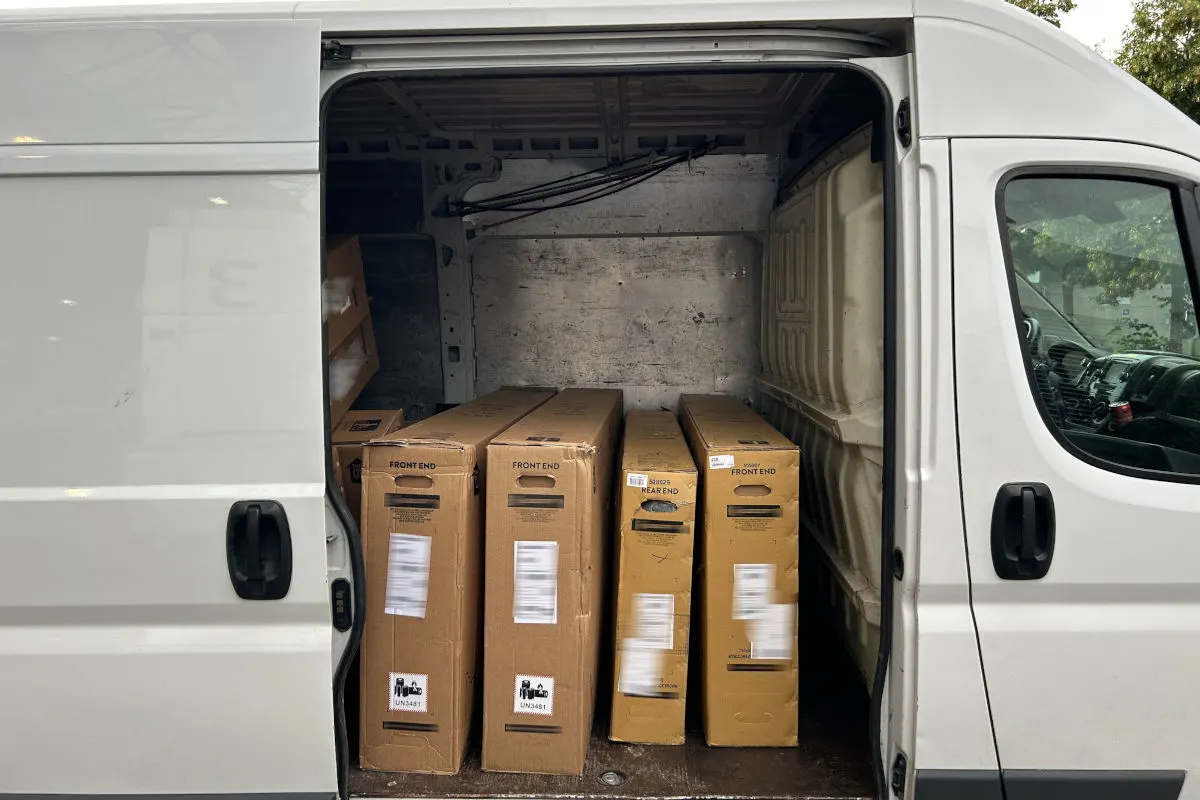 Ein weißer Transporter mit geöffneter Seitentür zeigt mehrere große, braune Kartons im Laderaum. Der Hintergrund ist ein Parkplatz.