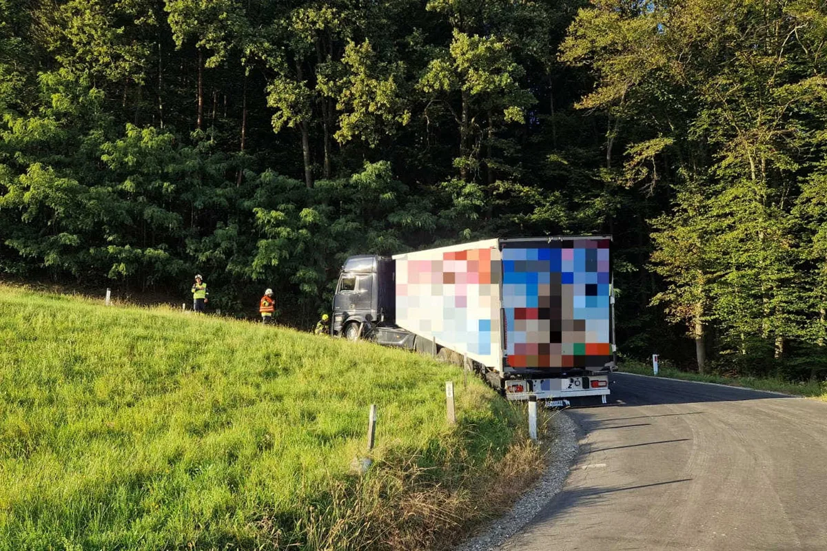 Ein schwarzer Lkw steckt in einer engen Kurve an einem Waldrand fest. Zwei Personen in Warnwesten stehen daneben auf der Wiese.
