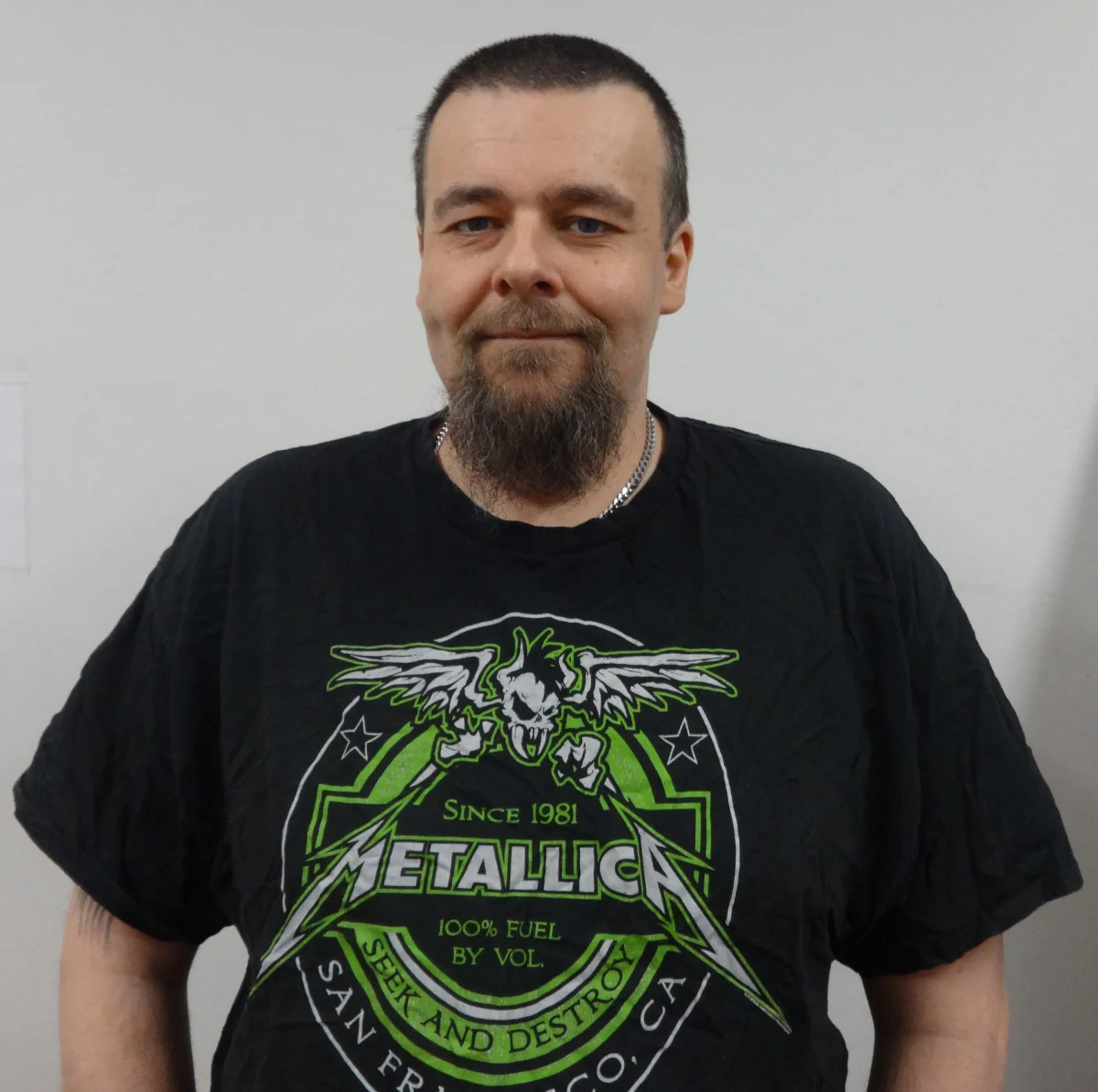 Ein Mann mit kurzem Haar und Bart trägt ein schwarzes T-Shirt mit einem auffälligen Metallica-Design in grüner Farbe. Der Hintergrund ist neutral, und der Fokus liegt auf seinem Oberkörper.