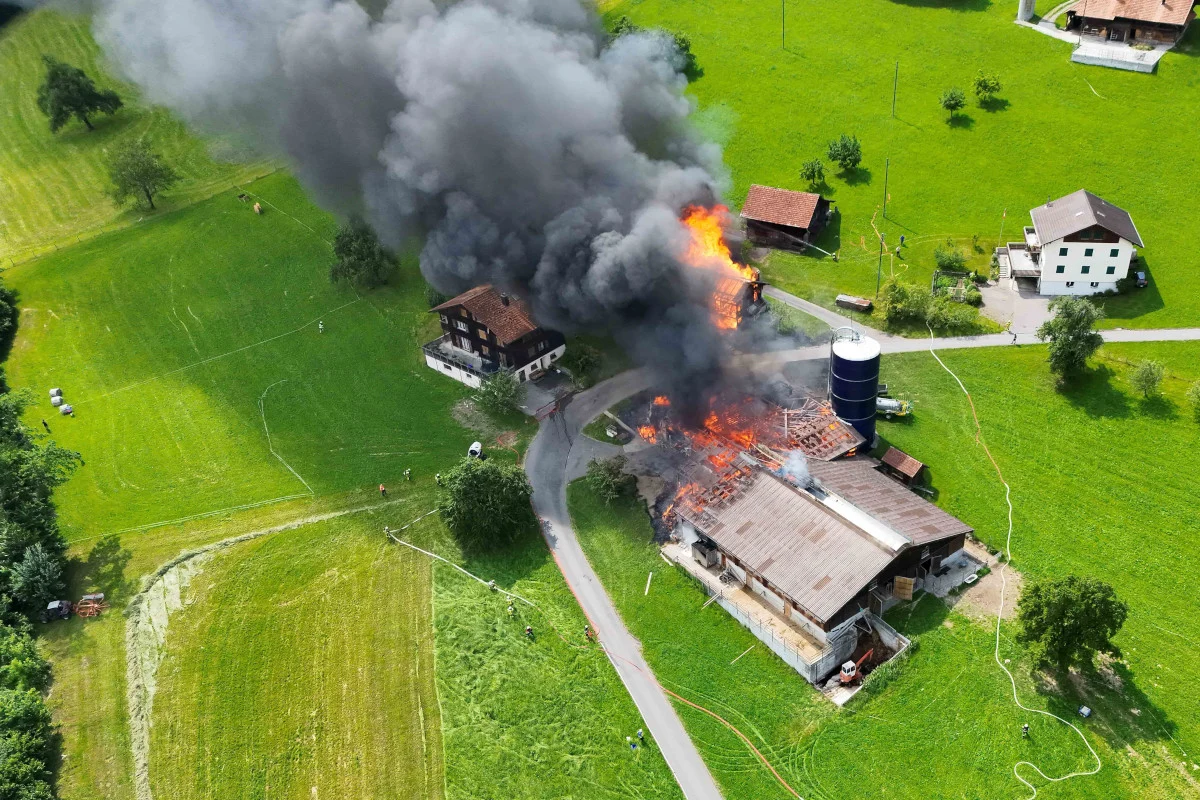 Ein Bauernhof steht in Flammen, dichter schwarzer Rauch steigt auf. Umgebung ist ländlich mit grünen Feldern.