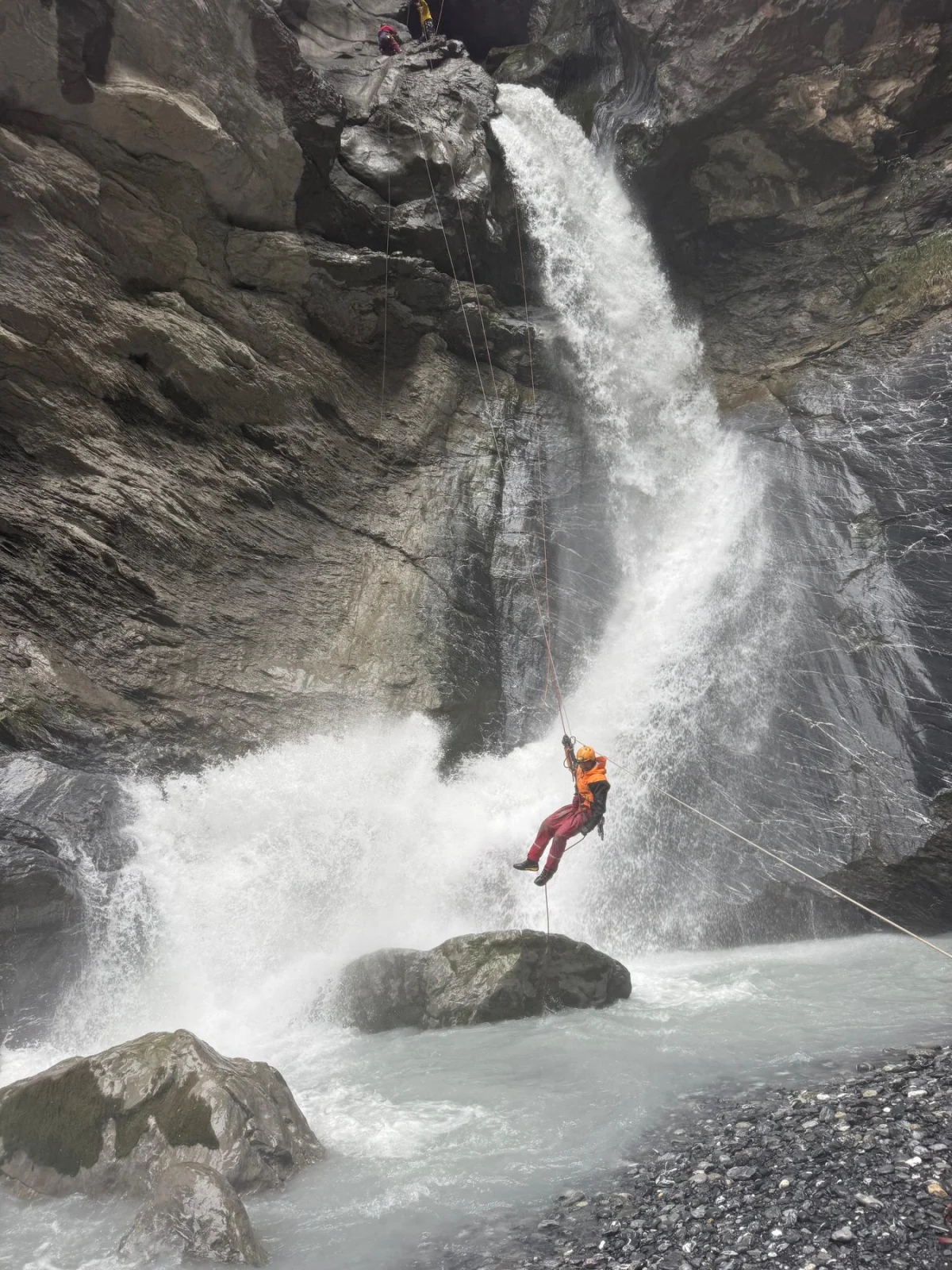 Eine Person in Rettungsausrüstung seilt sich an einem steilen Felsen neben einem Wasserfall ab. Zwei weitere Personen sind oben am Fels sichtbar.