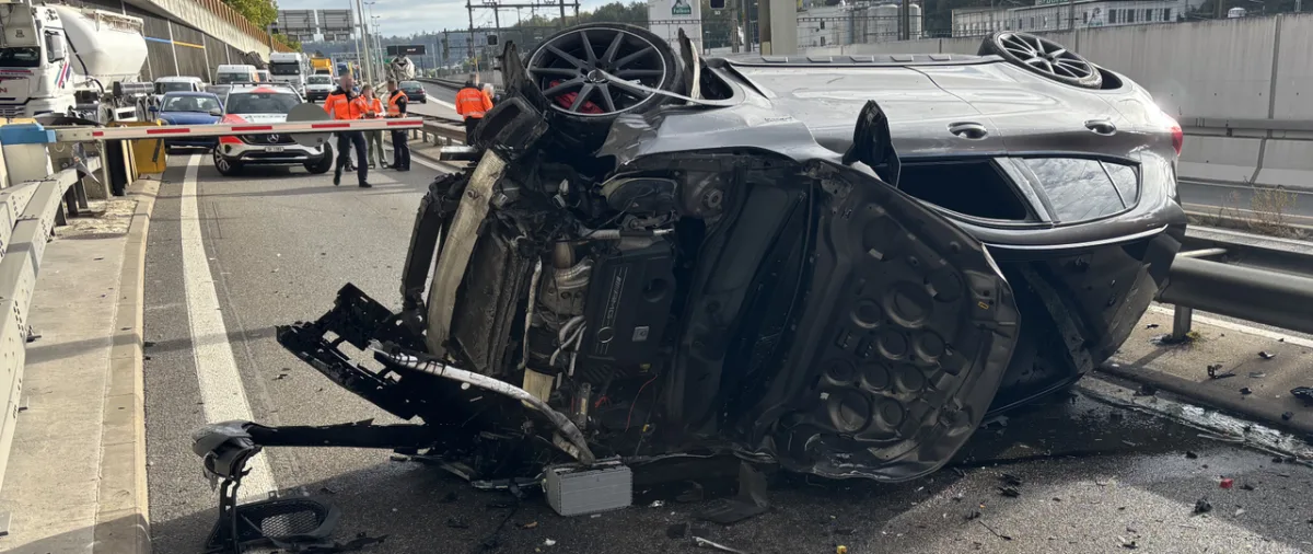 Ein silberner Sportwagen liegt verunfallt auf der Seite auf einer mehrspurigen Autobahn. Im Hintergrund sind Personen in Warnwesten zu sehen.