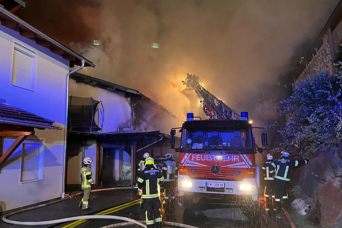 Die Feuerwehr ist mit einem Leiterfahrzeug vor einem brennenden Gebäude im Einsatz. Rauch steigt aus dem Dach auf, während Feuerwehrleute am Boden agieren.