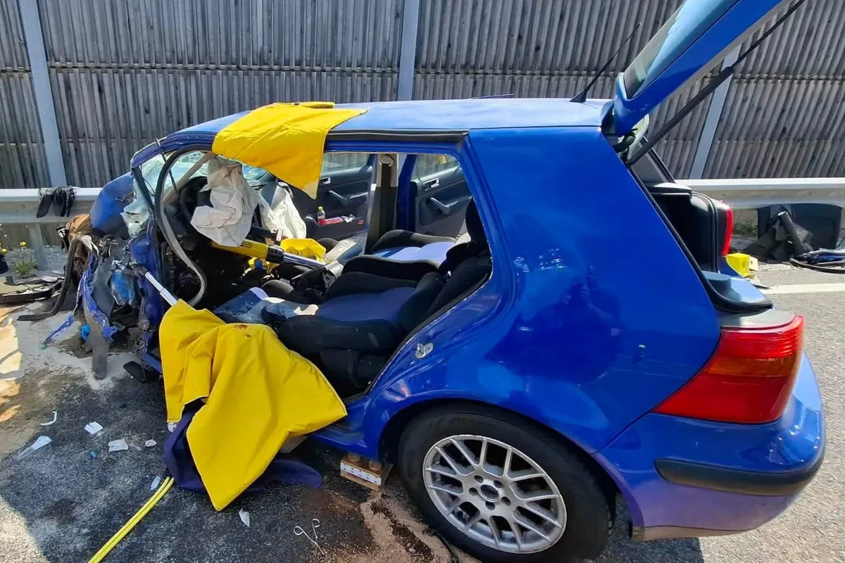 Ein schwer beschädigtes blaues Auto steht auf einer Autobahnspur. Der Kofferraum ist geöffnet und Trümmer liegen um das Fahrzeug verstreut.