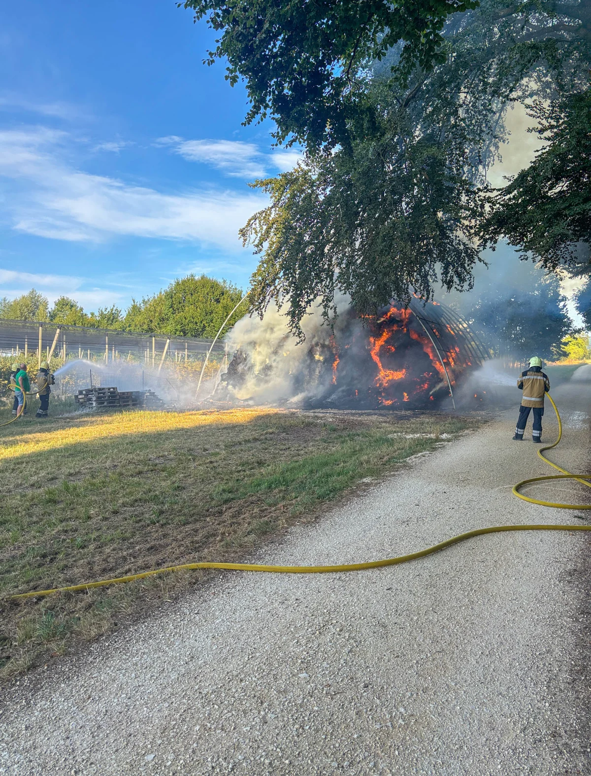 Ein brennendes, mit Flammen überzogenes Gebäude wird von Feuerwehrleuten mit Wasserschläuchen in einer ländlichen Umgebung gelöscht. Rauch steigt auf.
