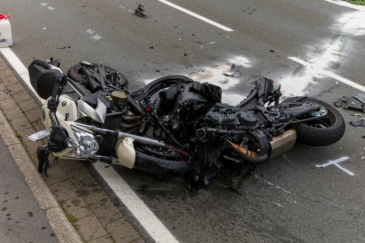 Zwei Motorräder liegen stark beschädigt auf einer nassen Fahrbahn. Um sie herum sind Trümmer und weiße Markierungen.