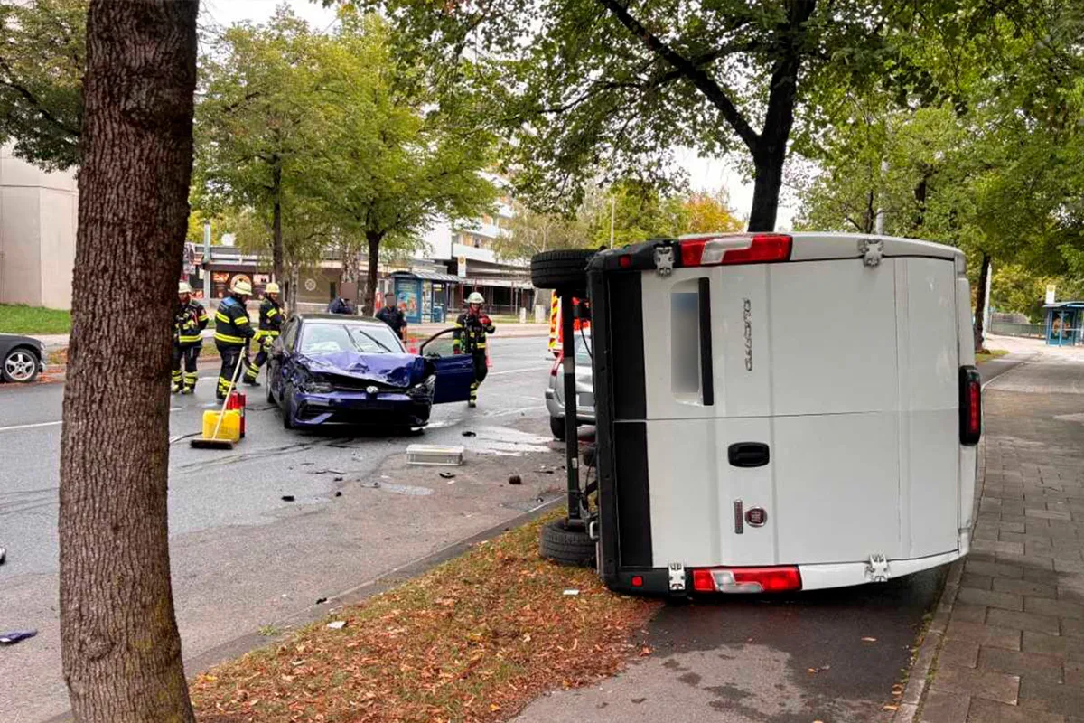 Ein weißer Van liegt auf der Seite, daneben ein beschädigtes blaues Auto. Feuerwehrleute sind vor Ort auf einer Stadtstraße zu sehen.