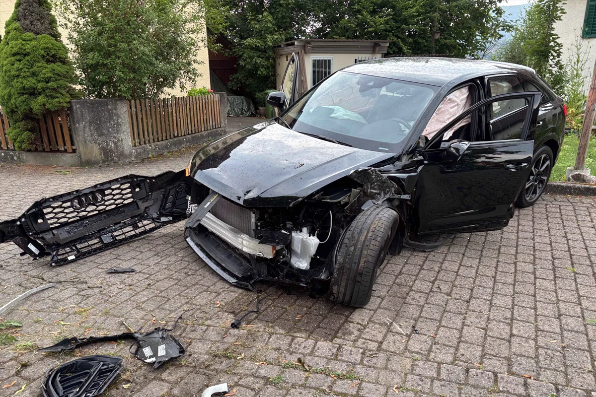 Ein stark beschädigtes schwarzes Auto steht auf einem gepflasterten Vorplatz. Der Stoßfänger liegt abgerissen daneben.