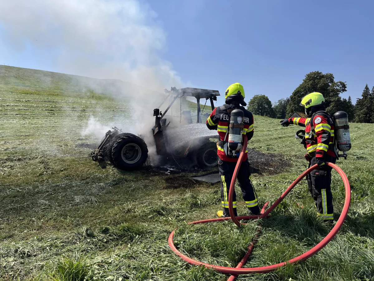Zwei Feuerwehrleute löschen einen brennenden Traktor auf einem Feld. Dicker Rauch steigt vom Traktor auf, während sie mit Schläuchen arbeiten.