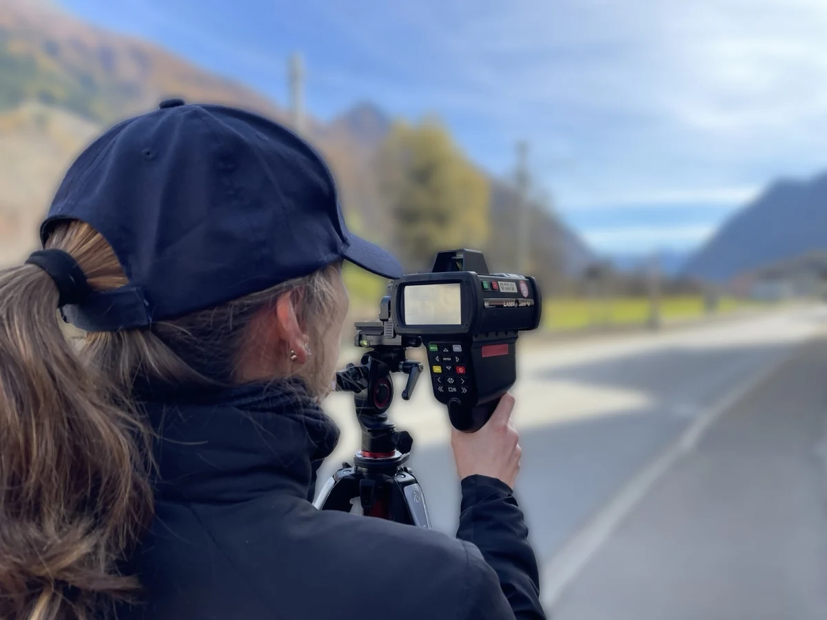 Eine Polizistin steht am Straßenrand und benutzt ein Lasermessgerät bei klarem Himmel. Im Hintergrund ist eine Landschaft mit Bergen zu sehen.