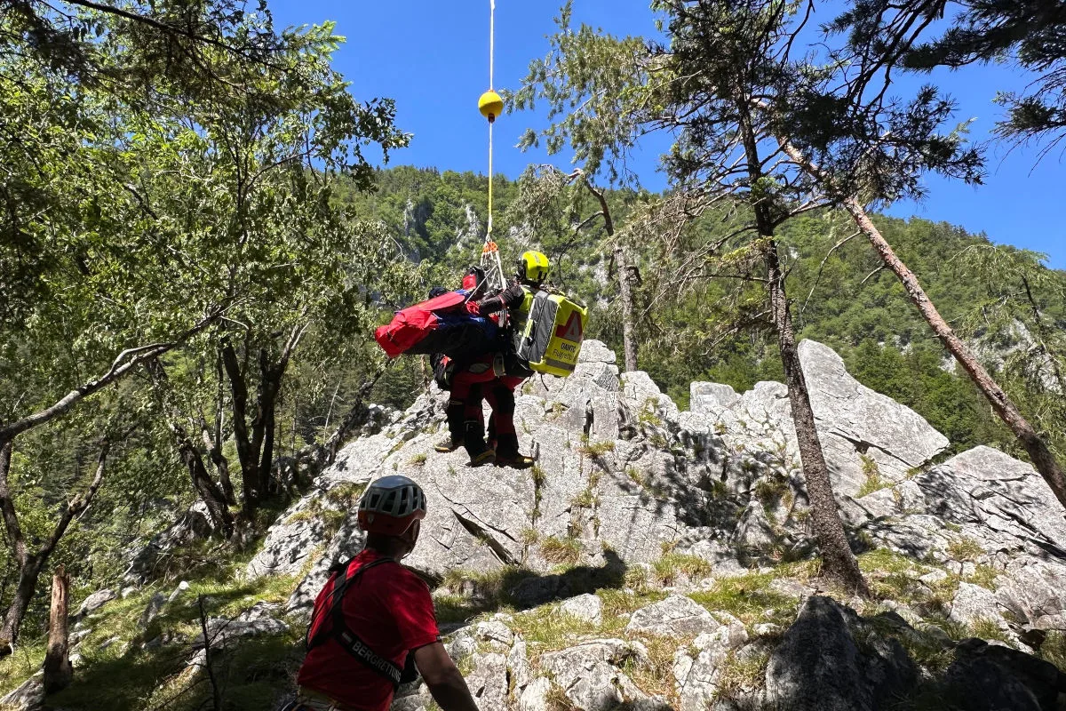 Ein Bergretter schwebt mit Ausrüstung an einem Seil über felsigem Gelände in einer bewaldeten Berglandschaft.