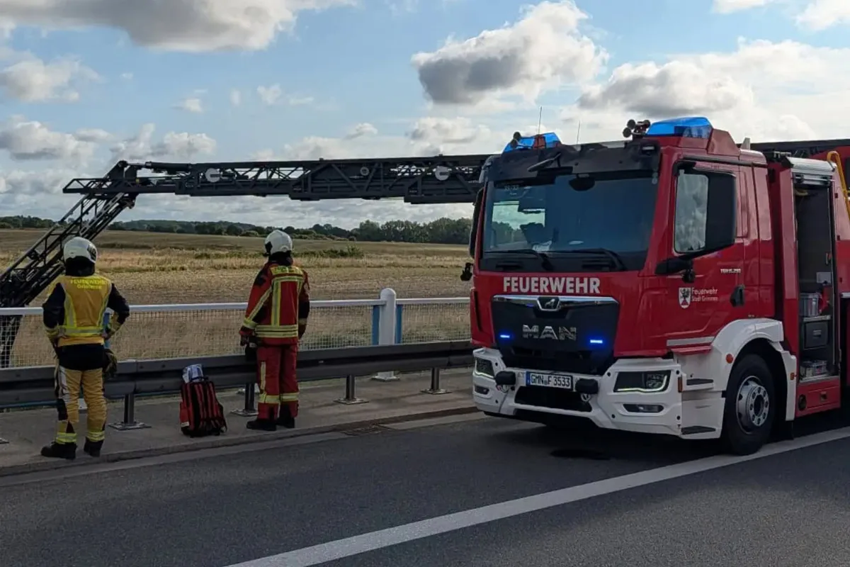 Ein rotes Feuerwehrfahrzeug steht auf einer Straße, flankiert von zwei Feuerwehrleuten in Einsatzkleidung. Im Hintergrund ist eine ländliche Landschaft zu sehen.