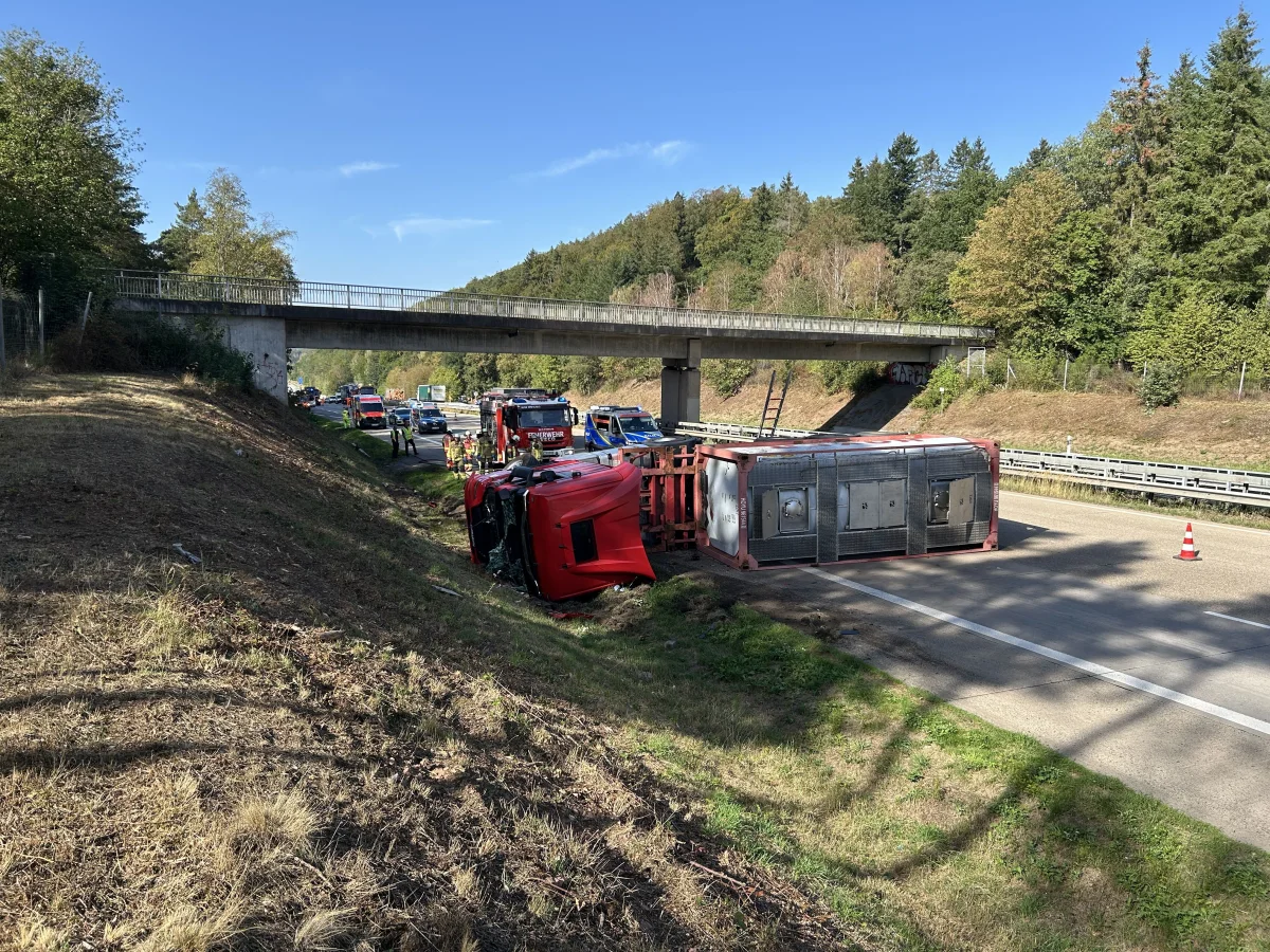 Ein roter Lkw liegt umgekippt auf der rechten Spur einer Autobahn, während Einsatzkräfte arbeiten.