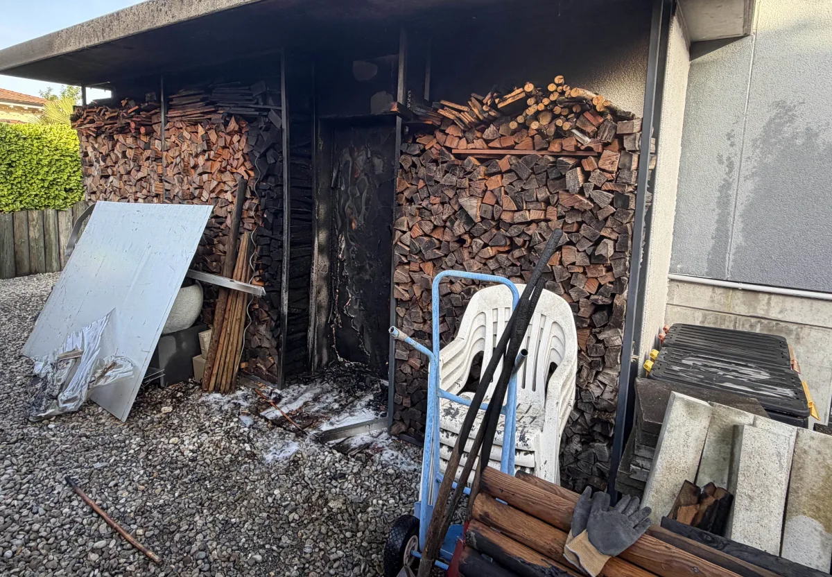 Ein verbranntes Regal mit Holzscheiten in einem Außenbereich zeigt Brandspuren. Daneben befindet sich abgelagerter Schutt.
