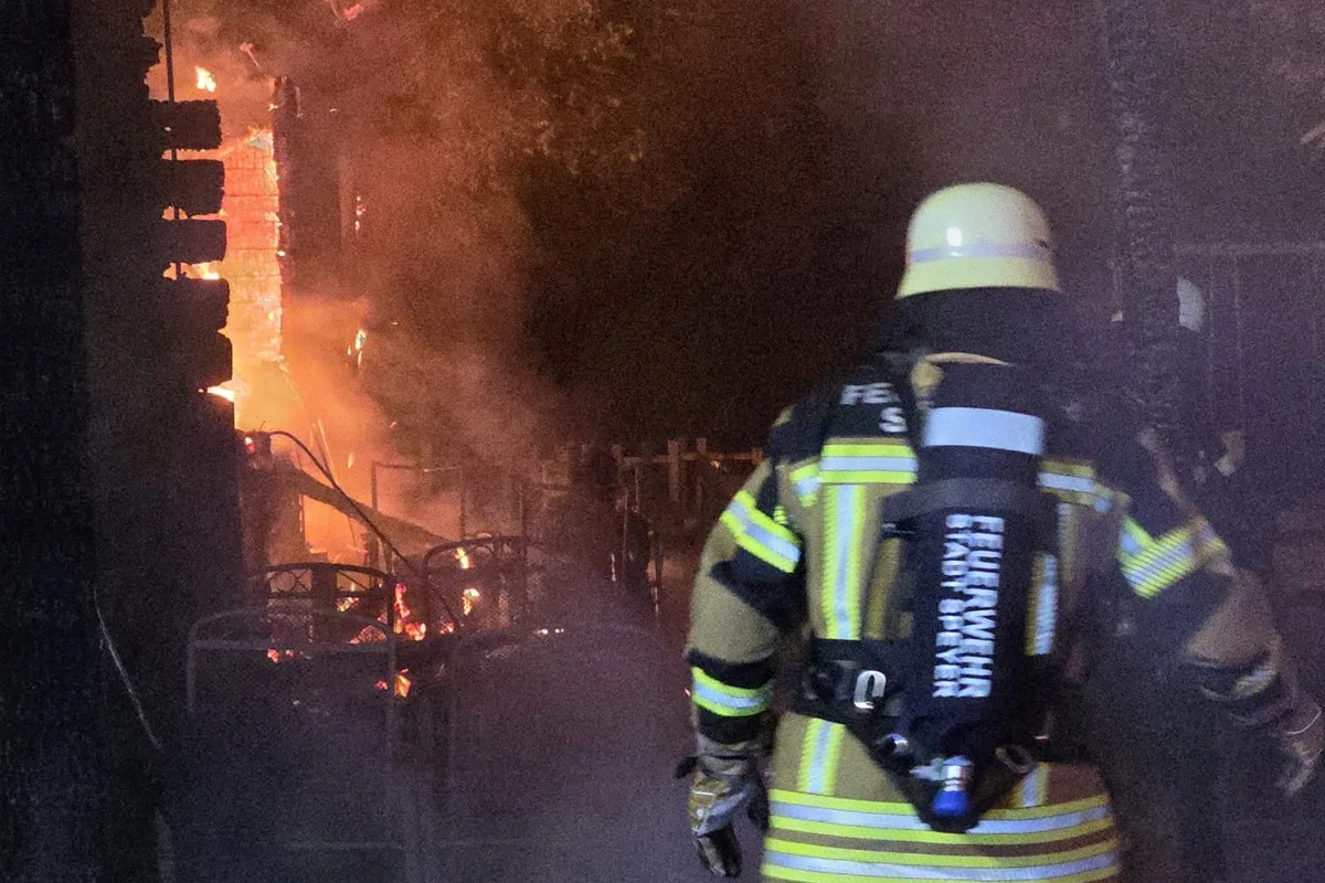 Ein Feuerwehrmann in voller Einsatzkleidung steht vor einem Gebäude, das in Flammen steht. Rauch und Flammen sind im Hintergrund sichtbar, es ist Nachtzeit.