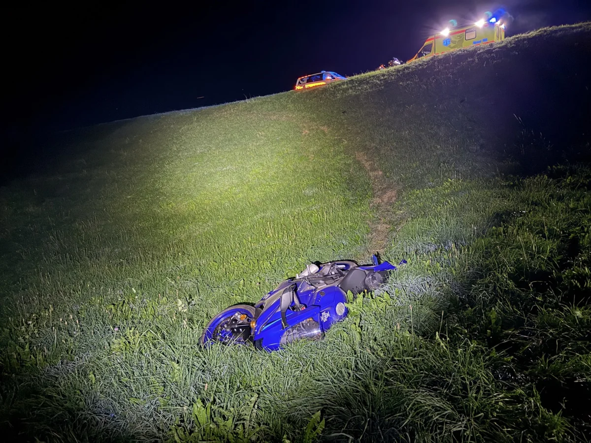 Ein blaues Motorrad liegt nachts auf einer Wiese, im Hintergrund ein beleuchtetes Einsatzfahrzeug auf der Straße.