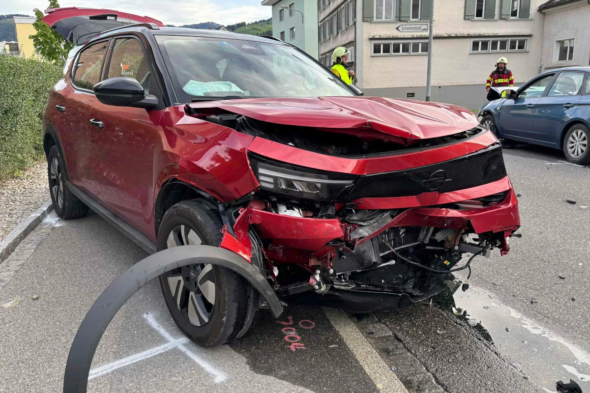 Ein roter SUV auf einer Straße zeigt erhebliche Frontschäden aus einem Unfall. Im Hintergrund stehen zwei Feuerwehrleute.