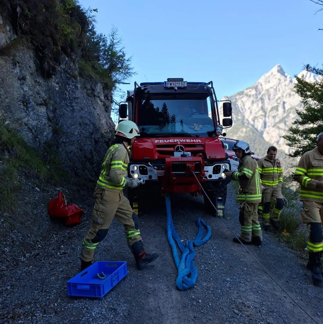 Ein rotes Feuerwehrfahrzeug steht auf einem schmalen Bergweg. Einsatzkräfte in Uniform bereiten ein blaues Seil vor.