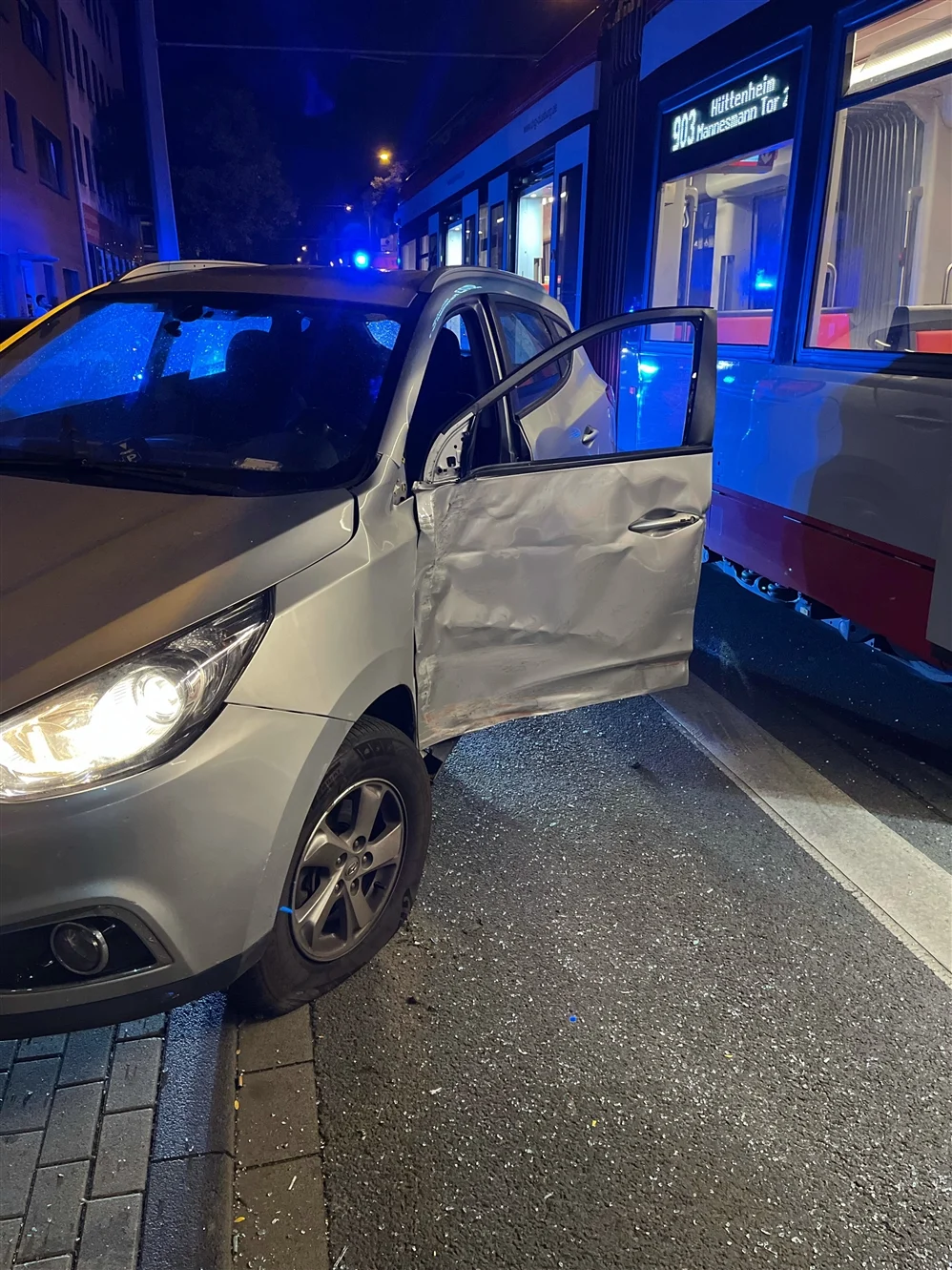 Bei Nacht ist ein silbernes Auto mit einer Straßenbahn kollidiert. Die Fahrertür des Autos ist stark beschädigt und offen.