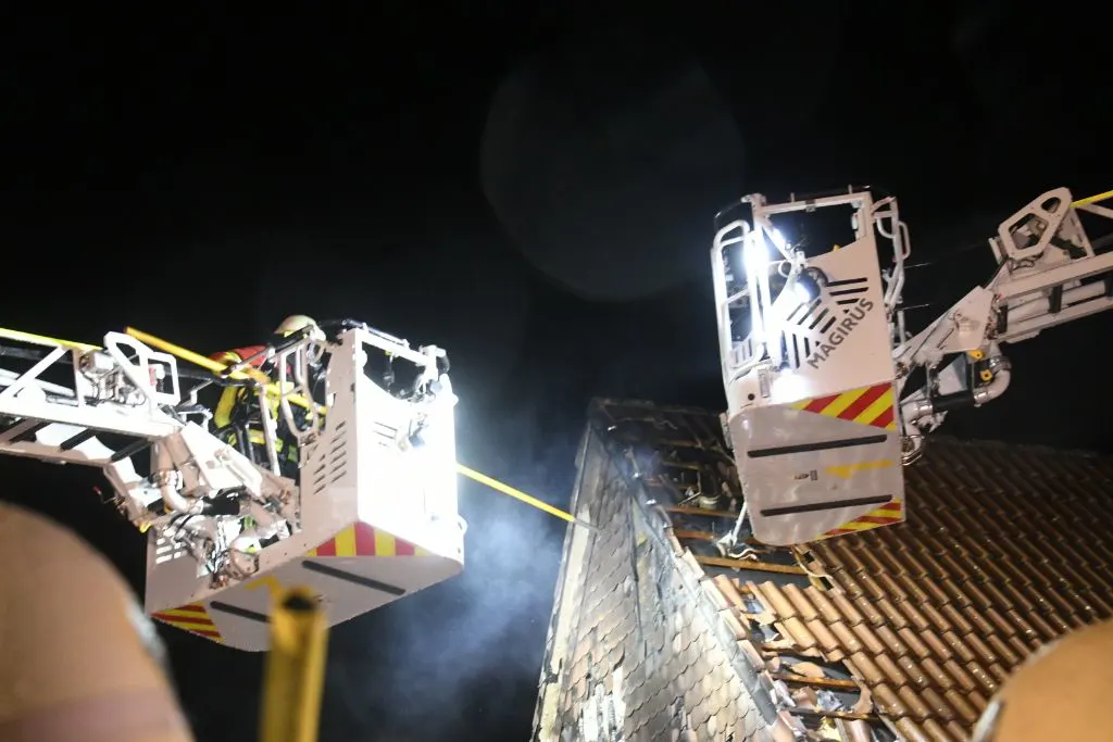 Zwei Feuerwehrarbeitskörbe halten Feuerwehrleute mit Schläuchen vor einem beschädigten Dach bei Nacht. Sicherheitslichter sind aktiv.