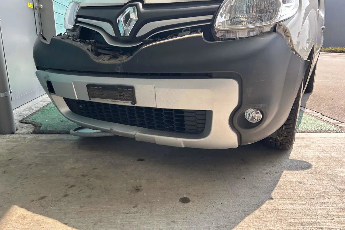 Ein weißer Lieferwagen mit sichtbaren Frontschäden und einer eingedrückten Stoßstange steht auf einem Parkplatz.