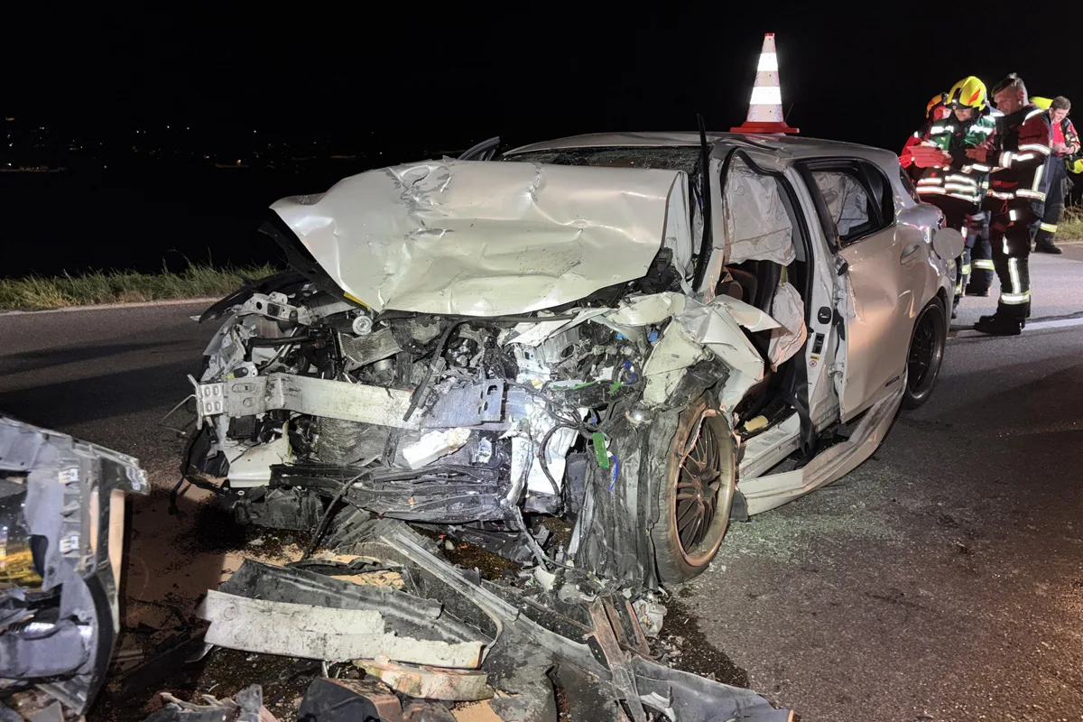 Ein stark demoliertes weißes Auto steht auf einer Straße bei Nacht. Im Hintergrund sind mehrere Einsatzkräfte zu sehen.