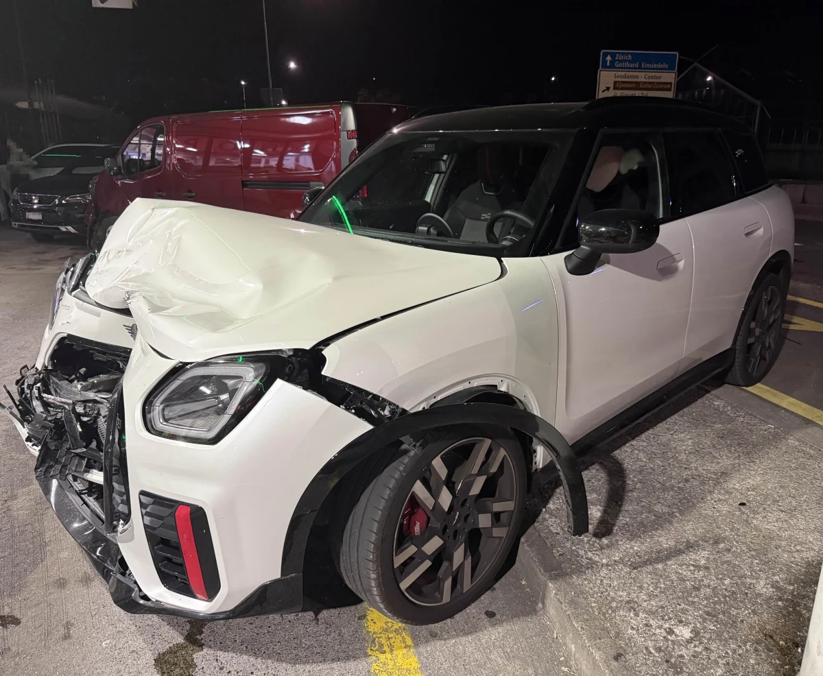 Ein weißer Mini Cooper steht mit zerstörter Frontpartie auf einem Parkplatz. Es ist Nacht, und der Schaden deutet auf einen heftigen Aufprall hin.