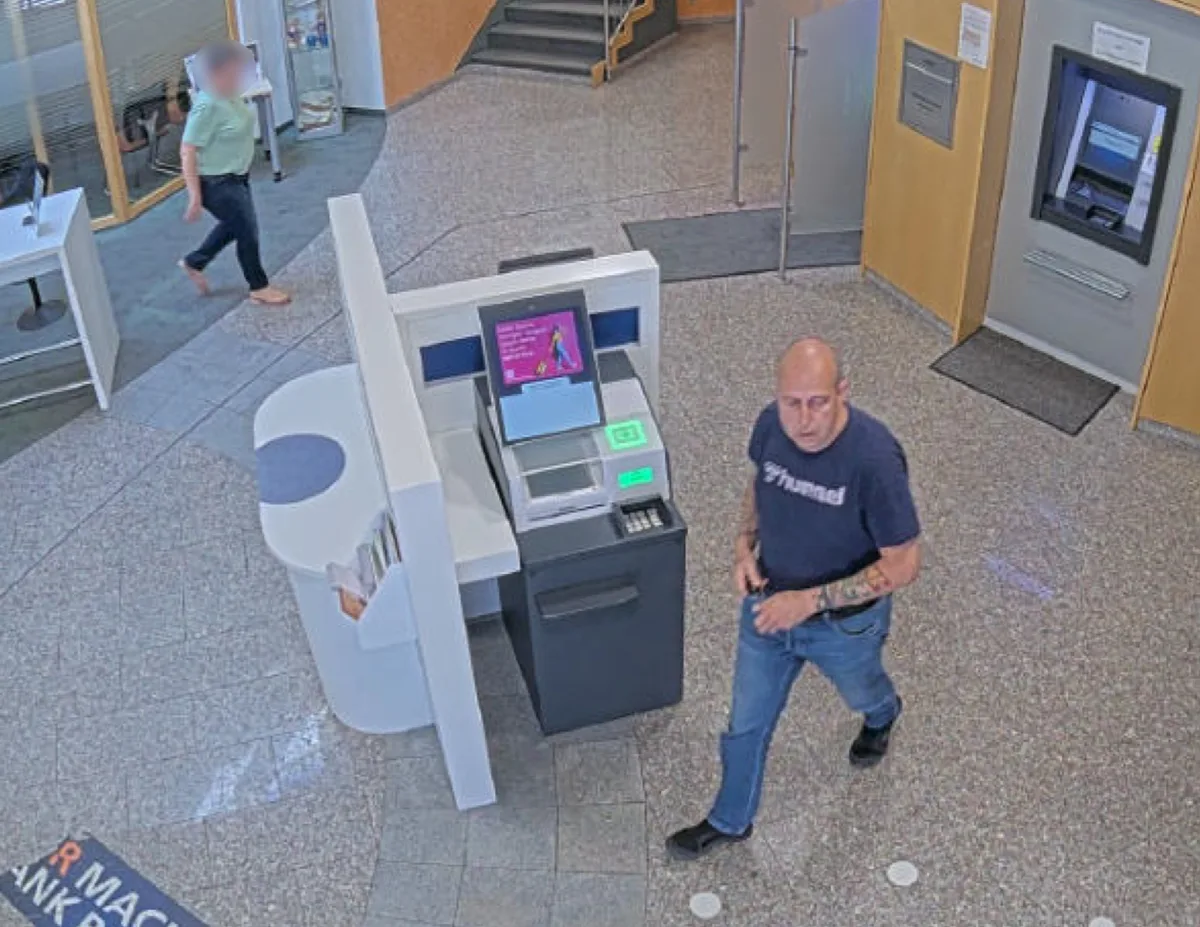 Das Bild zeigt einen Mann in einem Bankfoyer neben einem Geldautomaten, aufgenommen von einer Überwachungskamera.