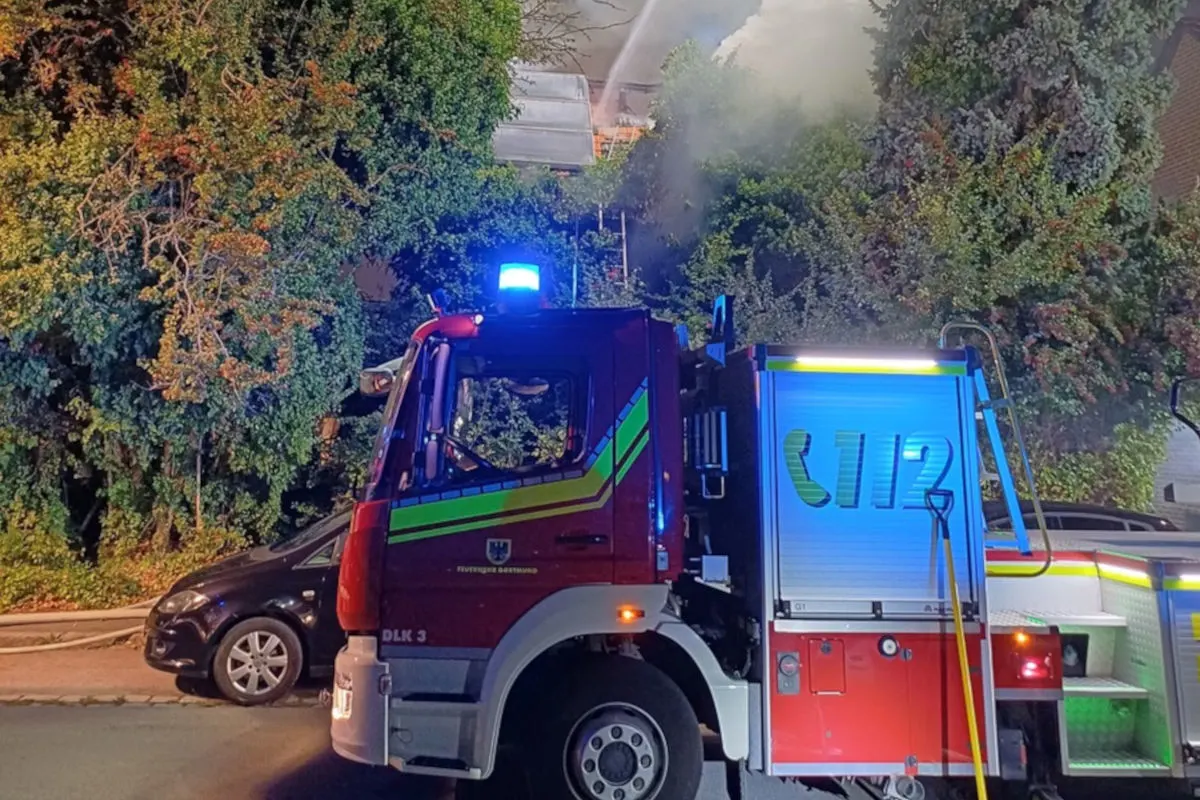 Ein rotes Feuerwehrfahrzeug steht vor einem brennenden Gebäude, aus dem Rauch aufsteigt. Es ist Nacht.