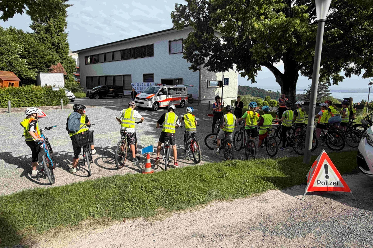 Eine Gruppe von Jugendlichen in leuchtend gelben Sicherheitswesten steht mit Fahrrädern auf einem Platz vor einem Gebäude. Ein Polizeifahrradschild ist im Vordergrund zu sehen und ein Einsatzfahrzeug ist im Hintergrund geparkt.