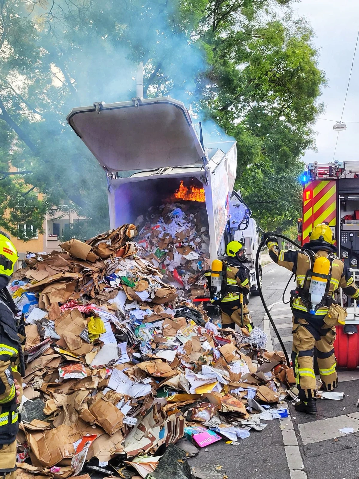 Ein Müllwagen steht in Flammen, während mehrere Feuerwehrleute das Feuer bekämpfen. Rauch steigt auf und Papiermüll liegt verstreut auf der Straße.