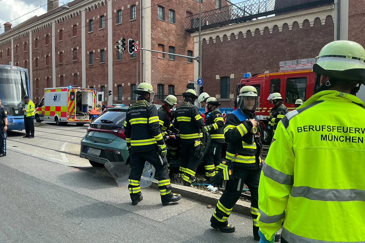 Mehrere Feuerwehrleute und ein Rettungswagen sind bei einem Einsatz auf einer Stadtstraße tätig. Ein Unfallfahrzeug ist im Hintergrund sichtbar.
