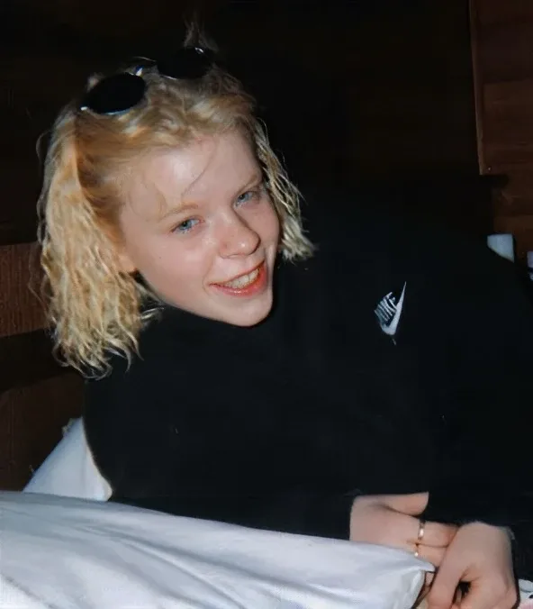 Eine blonde Person liegt lächelnd auf einem hellen Untergrund, trägt einen dunklen Pullover und eine Sonnenbrille.