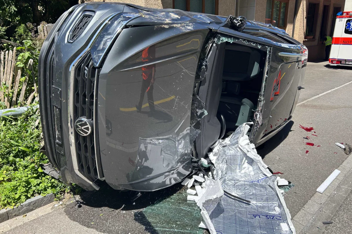 Ein schwarzes Auto liegt seitlich auf der Straße; die Front ist stark beschädigt, und das Glas der Fenster ist gebrochen.