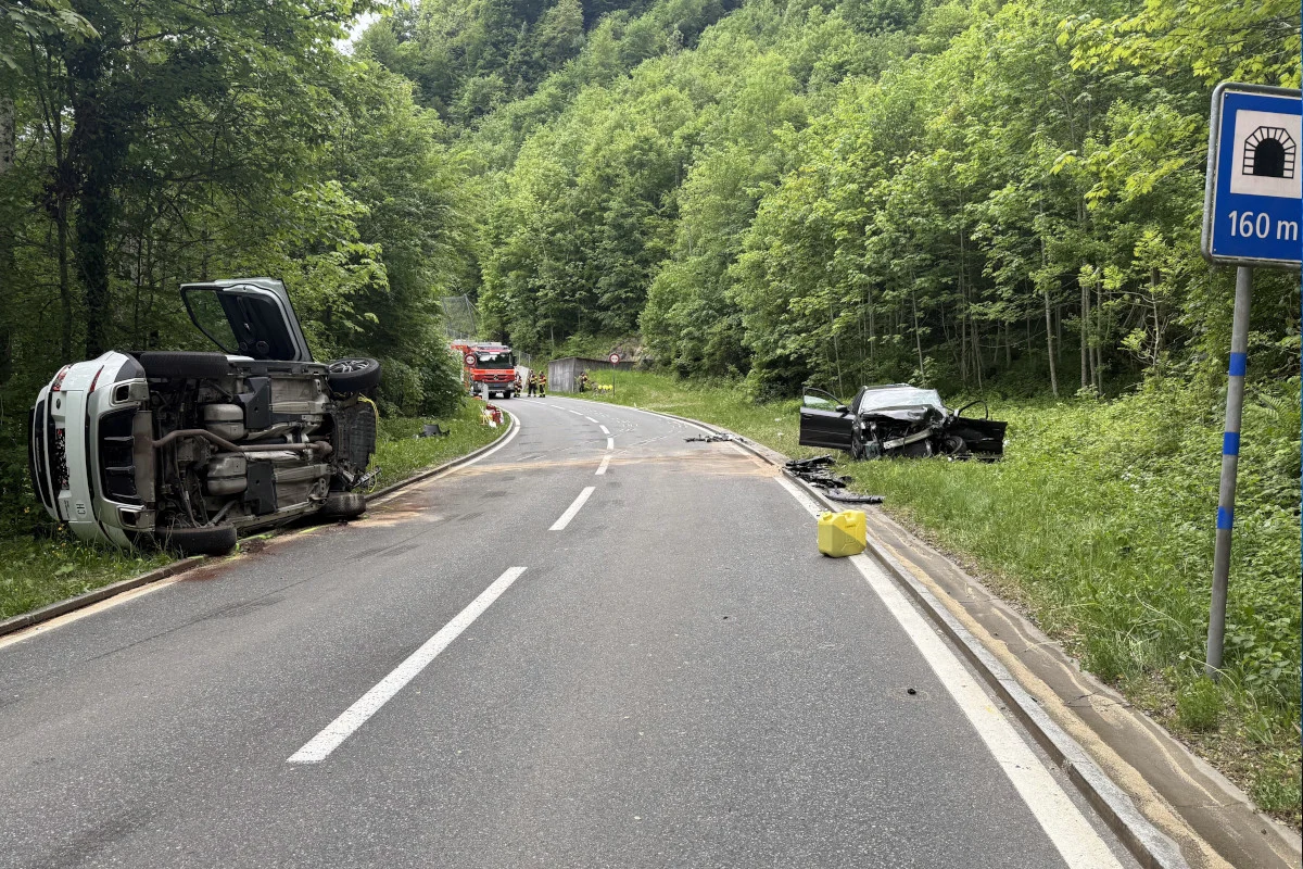 Ein Auto liegt auf der Seite im Gras, während das andere stark beschädigt am Straßenrand steht. Ein Feuerwehrfahrzeug und Rettungskräfte sind im Hintergrund zu sehen.