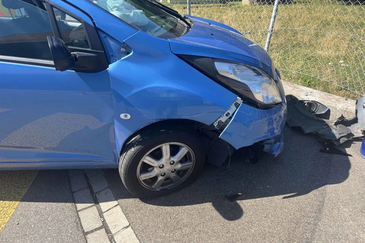 Ein blaues Auto steht am Straßenrand mit deutlichen Frontschäden und einem eingedrückten Kotflügel. Im Hintergrund ist ein Maschendrahtzaun sichtbar.