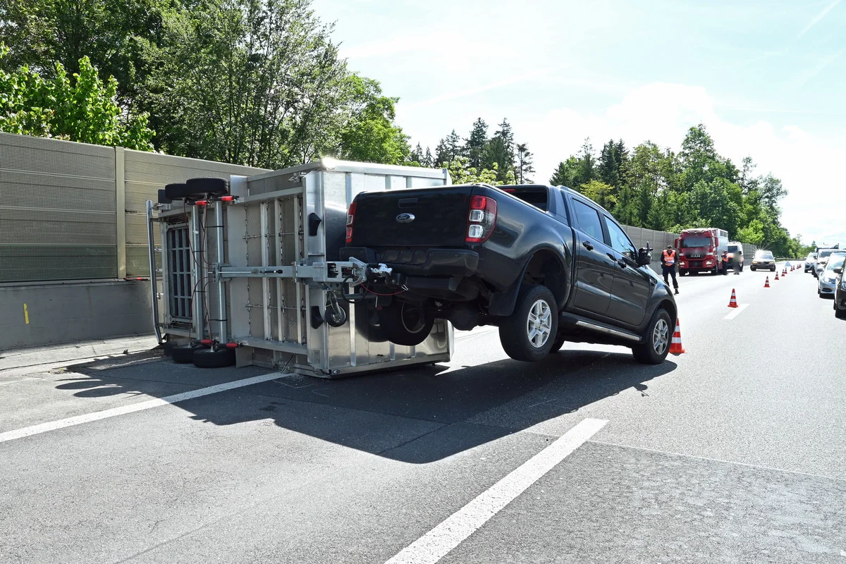 Ein schwarzer Pickup steht hinter einem umgekippten Anhänger auf der Autobahn. Verkehrskegel sind aufgestellt.
