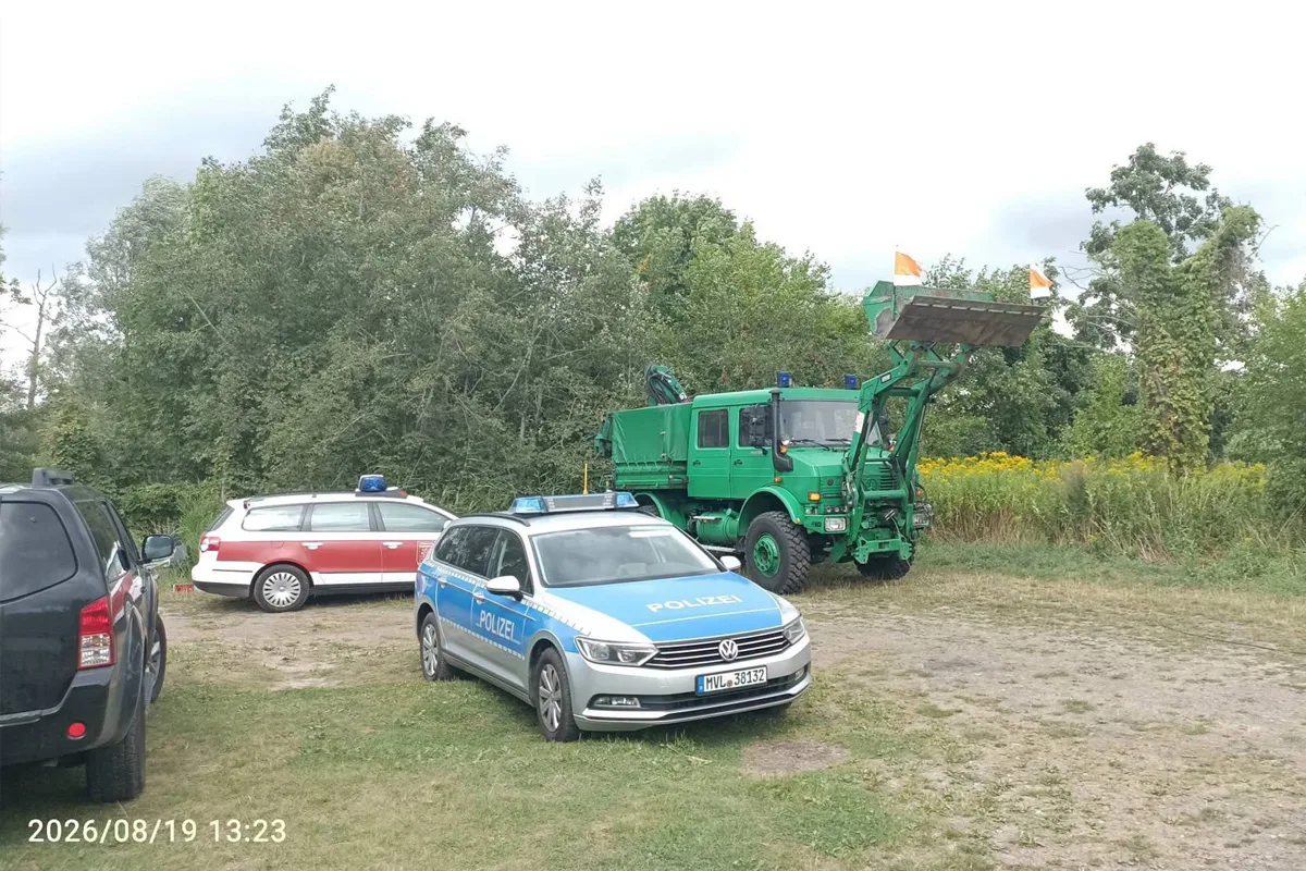 Ein deutsches Polizeiauto steht vor einem großen grünen Spezialfahrzeug neben einem Waldstück, während weitere Autos im Hintergrund zu sehen sind.