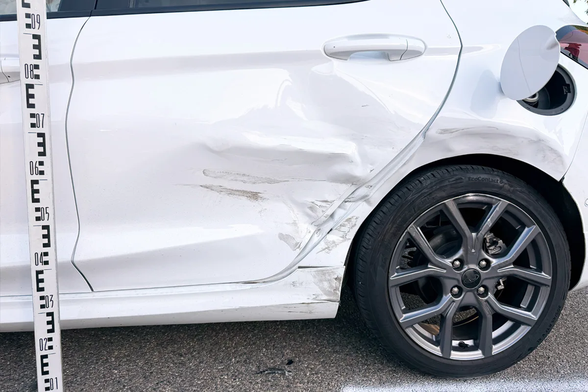 Die rechte Autotür eines weißen Fahrzeugs zeigt Dellen und Kratzer neben einer geparkten Felge. Ein Messstab steht daneben.