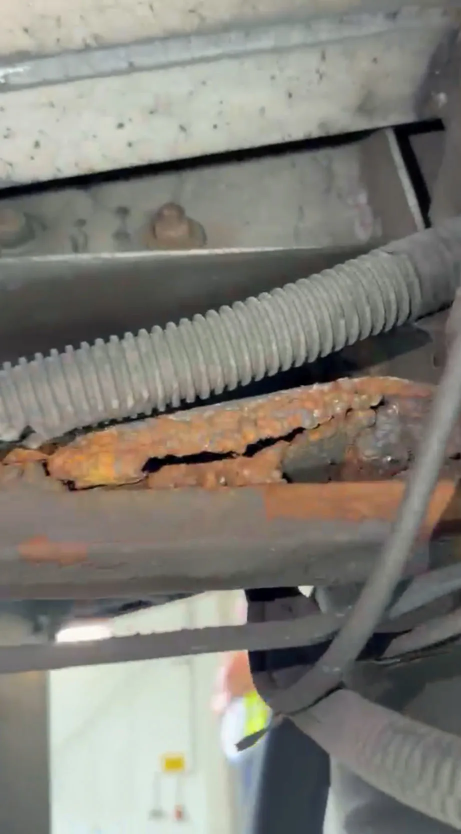 Ein stark verrosteter Metallträger unter einem Fahrzeug zeigt erhebliche strukturelle Schäden mit rissiger Oberfläche. Im Hintergrund sind verschwommen weitere Teile sichtbar.