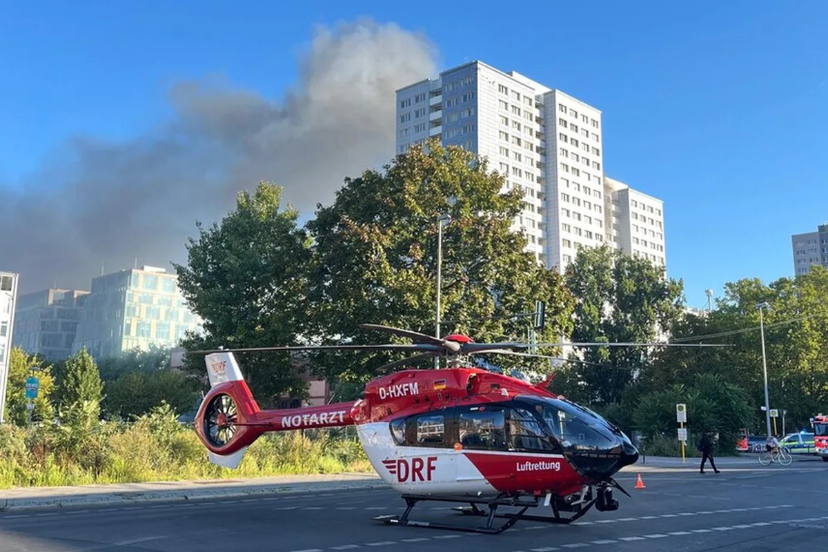Ein Rettungshubschrauber der DRF steht auf einer Straße in einer städtischen Umgebung. Im Hintergrund sind hohe Gebäude und Rauch sichtbar.