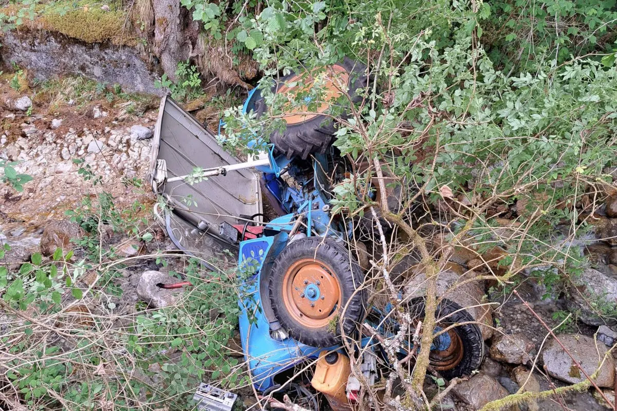 Ein blauer Traktor liegt umgestürzt im Bachbett, umgeben von Pflanzen und Bäumen. Teile des Traktors sind beschädigt.