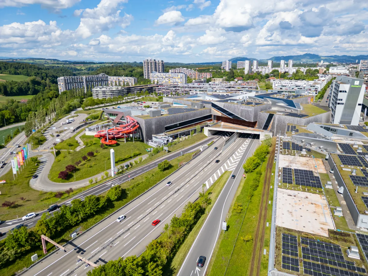 Das Bild zeigt ein modernes Einkaufszentrum im Vordergrund mit einer gut befahrenen Autobahn. Im Hintergrund sind Wohngebäude und ein bewaldeter Hügel zu sehen, bei sonnigem Wetter.