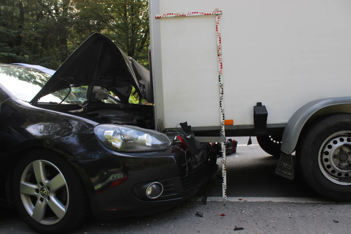 Ein schwarzer Pkw ist frontal mit einem weißen Anhänger kollidiert. Die Motorhaube des Autos ist geöffnet und es scheint beschädigt.