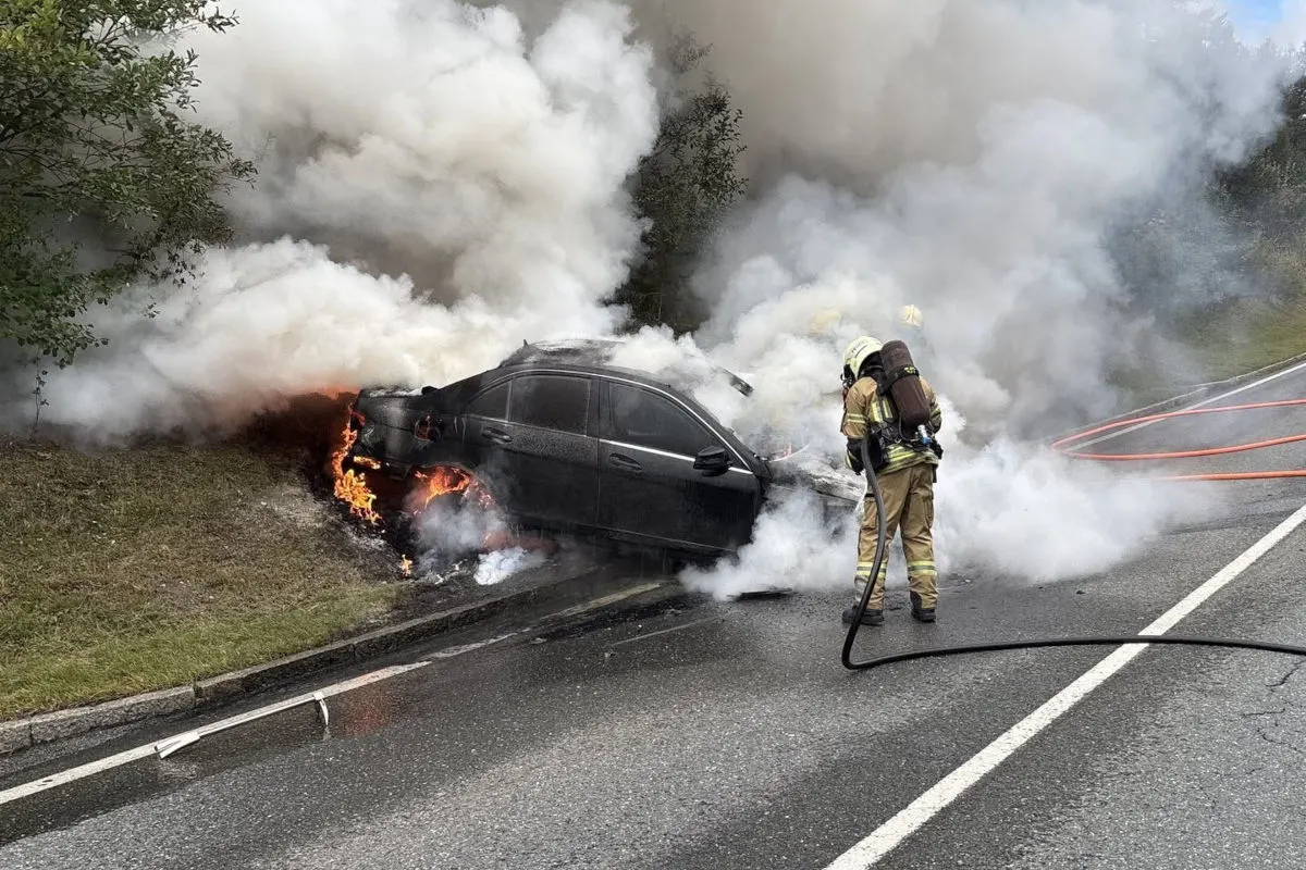 Ein Auto steht in Flammen am Straßenrand, während ein Feuerwehrmann den Brand löscht. Dichte Rauchwolken steigen auf.